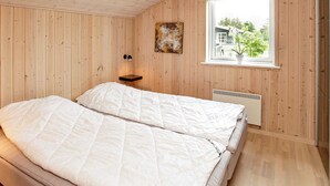 Et soveværelse med trævægge, en seng med hvide sengetæpper, et lille natbord med to lamper, en radiator og et vindue med udsigt til grønne omgivelser.