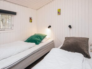 A room with two beds, one with a green pillow and the other with a brown pillow. There is a window on the left and two wall-mounted lamps.