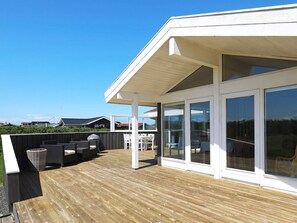 A covered outdoor deck with a wooden floor, white furniture, and large sliding glass doors leading to a covered patio area. - Spacious Holiday Home in Jutland With Sauna (Lokken)