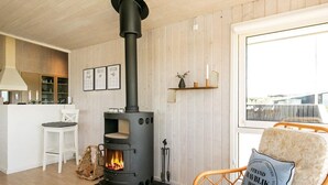 Une pièce chaleureuse avec un poêle à bois, un coin repas avec une chaise blanche et une chaise en osier avec un coussin.