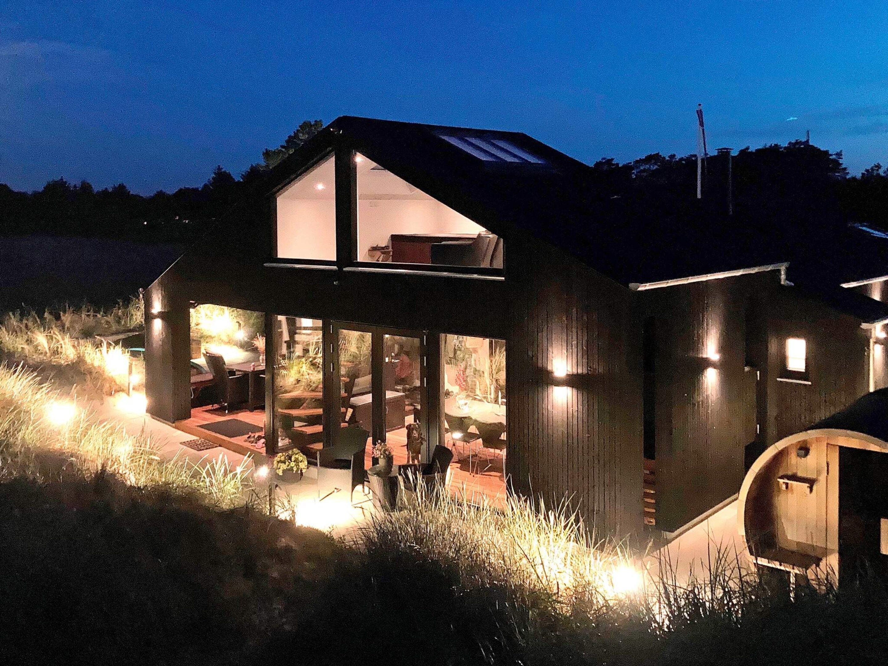 Ein modernes Haus mit einem großen Glasfenster, einer Holzdielen-Terrasse und Außenbeleuchtung.