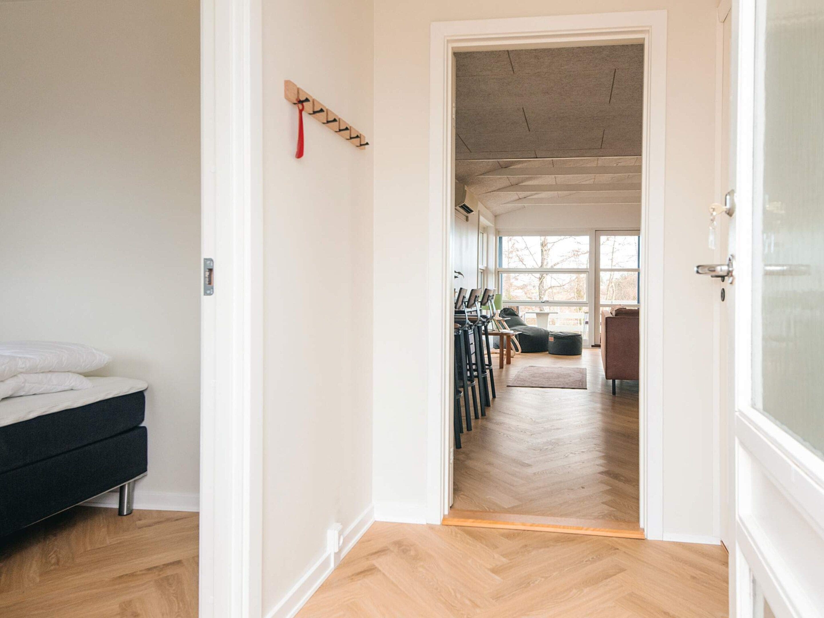 A room with a bed, a wooden floor, a door leading to another room with a sofa and chairs.