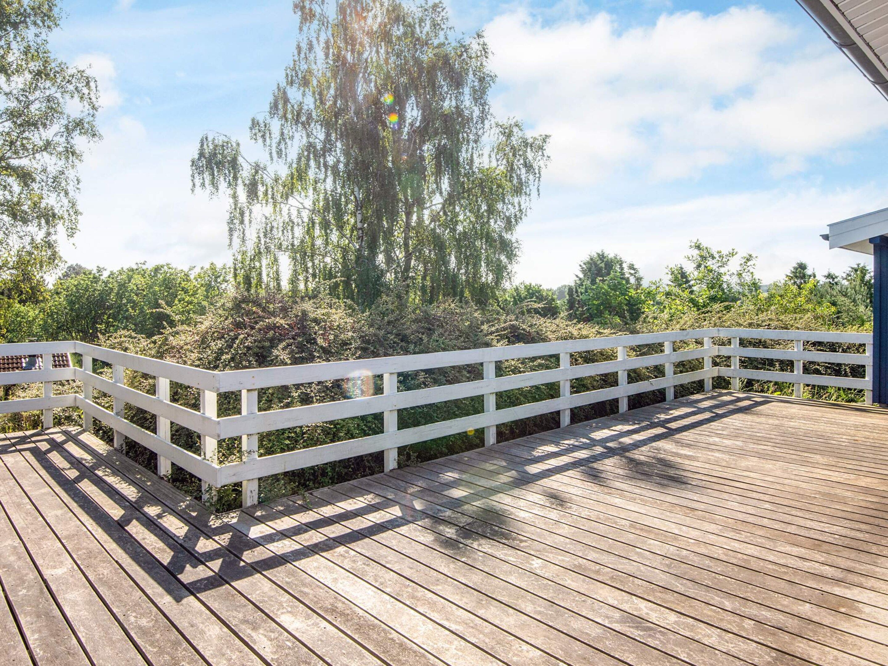 A wooden deck with white railings, surrounded by greenery and trees.