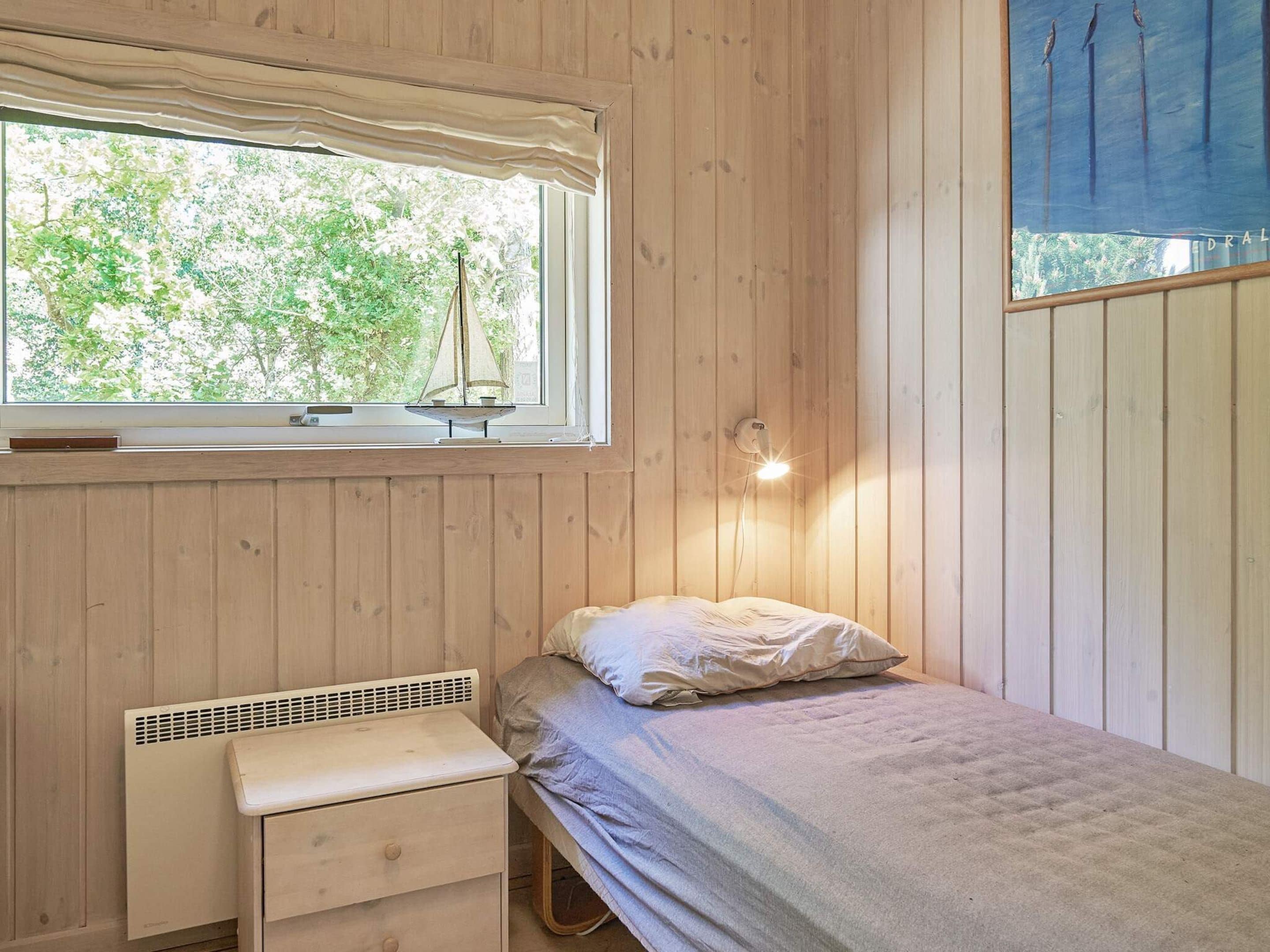 A wooden room with a bed, a small table, a window with a view of trees, and a wall-mounted light.