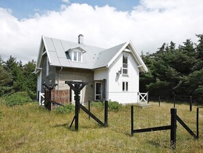 Une maison blanche avec un toit à pignon, entourée d’une clôture et de verdure.