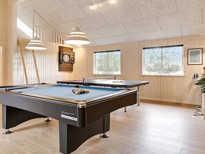 A pool table with a dartboard and billiard balls, wooden floors, and large windows.
