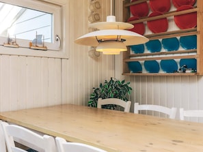 Une table à manger en bois, entourée de chaises blanches, un luminaire suspendu et une étagère murale ornée d’assiettes rouges et bleues.