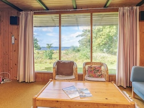 A room with two wicker chairs, a wooden table with magazines, and a view of the outdoors through large windows.