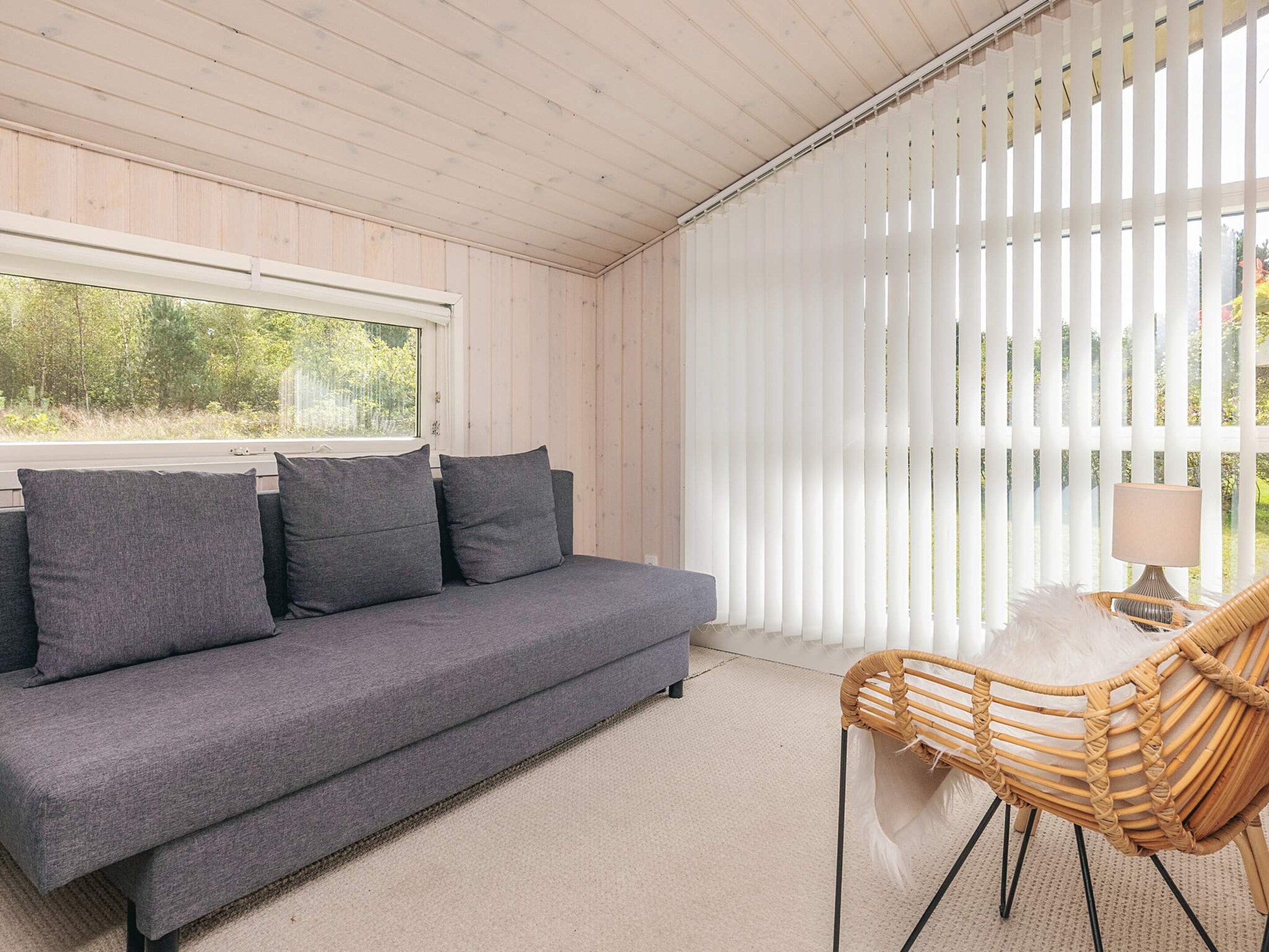 A room with a grey sofa, a wicker chair, and a window with white blinds.