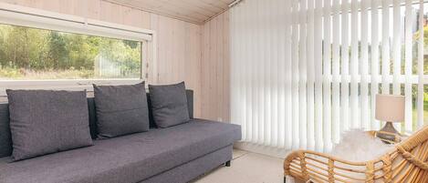 A room with a grey sofa, a wicker chair, and a window with white blinds.