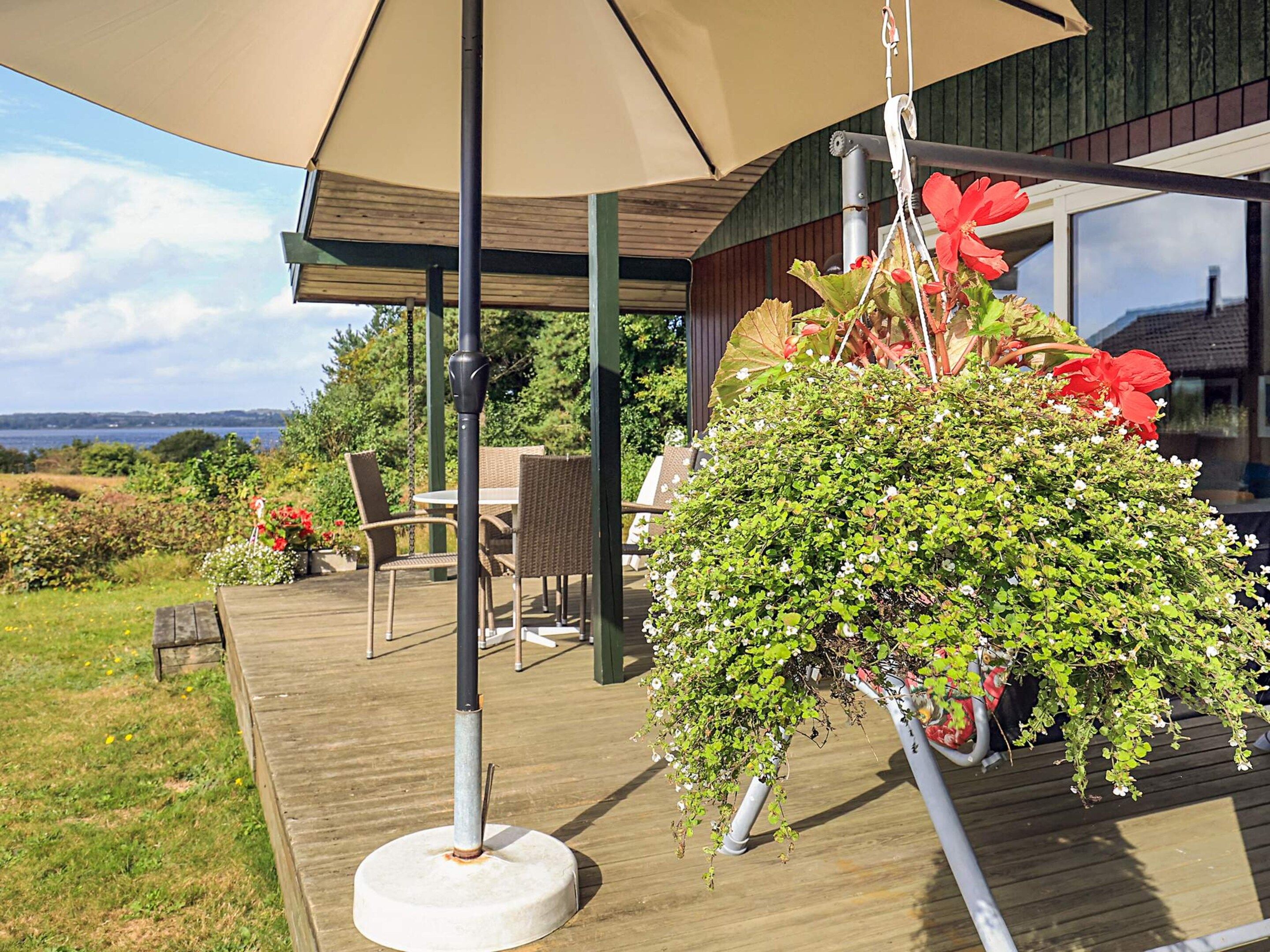 Una terraza de madera con mobiliario exterior, una maceta colgante con follaje y vistas del paisaje.