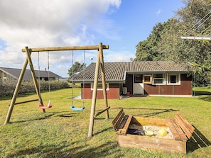 Un ensemble de balançoires en bois, un petit bâtiment doté d’un toit en tuiles et une structure de jeu en bois avec un banc.