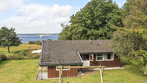 A small house with a tiled roof, a wooden deck, and a swing set in a grassy yard with a view of the sea. - 8 Person Holiday Home in Skals (Skals)