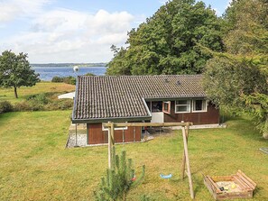 A small house with a tiled roof, a wooden deck, and a swing set in a grassy yard with a view of the sea.