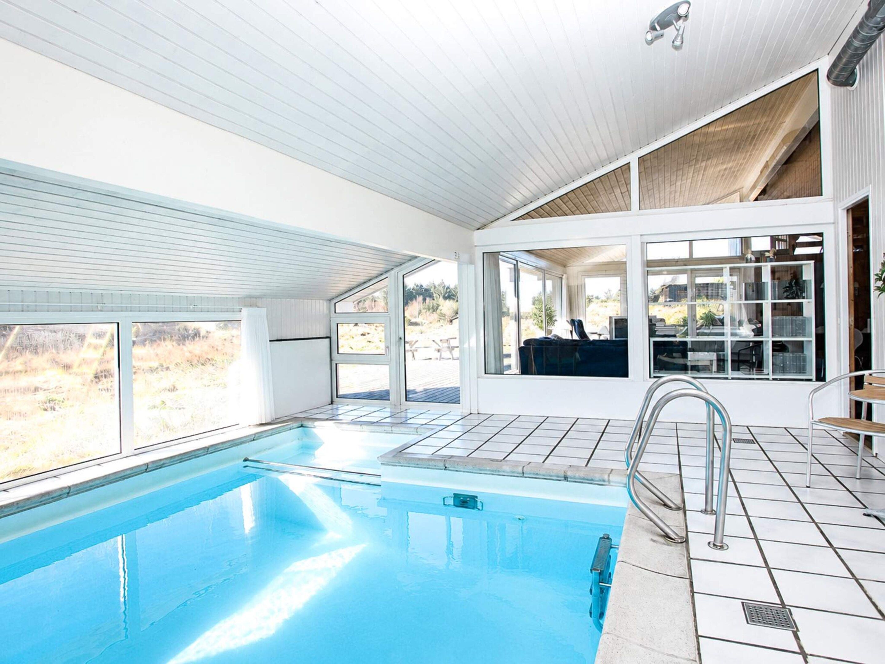 Una piscina interior moderna con paredes de vidrio transparente, piso de baldosas y vistas al exterior.