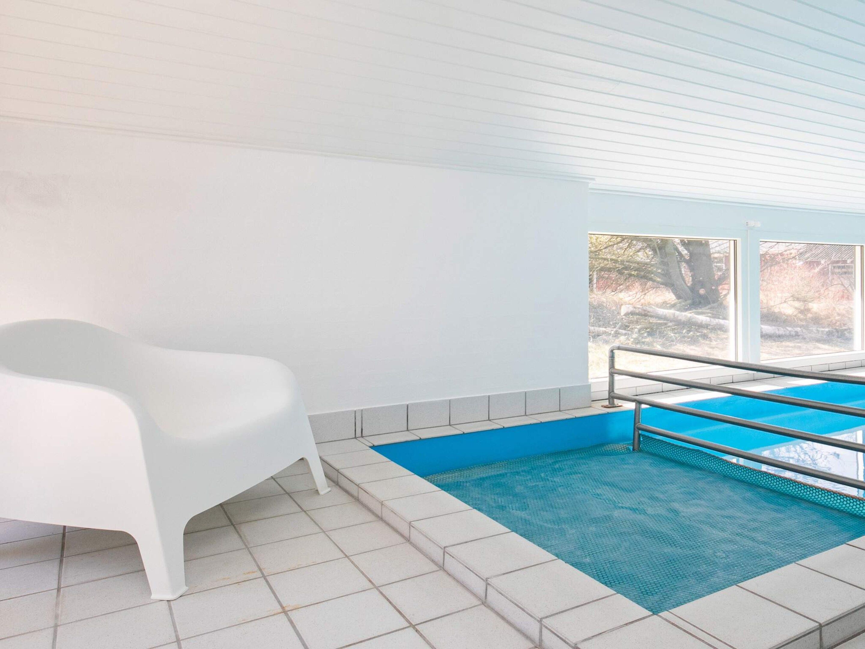 A swimming pool with a blue border, a white bench, and a window with a view of trees.
