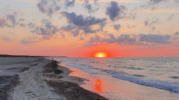 Ein Strand mit Sonnenuntergang, Wellen und einer Person, die mit einem Hund spazieren geht.