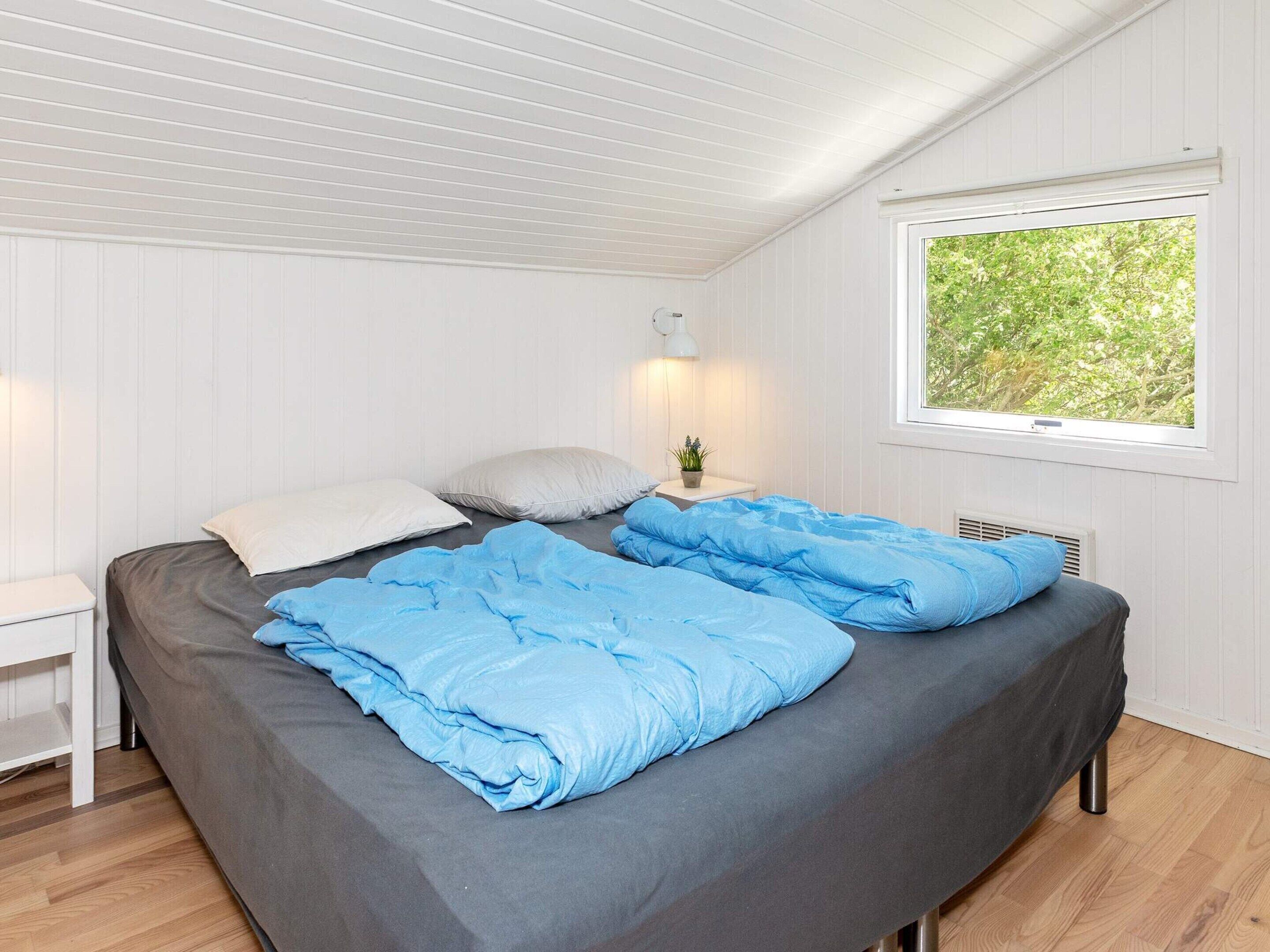 A single bed with a grey mattress and blue bedding, a bedside table with a lamp, and a window with a view of greenery.