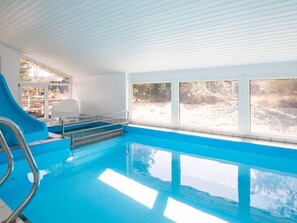 An indoor swimming pool with a slide, a sunlit room with large windows, and a view of a wooded area.