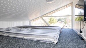 Quarto com teto inclinado, cama com roupa de cama listrada, TV fixada na parede e janela com vista para o exterior.