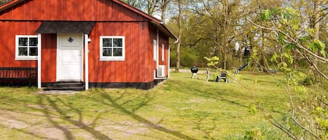 A red wooden cabin with a white door and windows, a chimney, and a grassy yard with a barbecue grill.