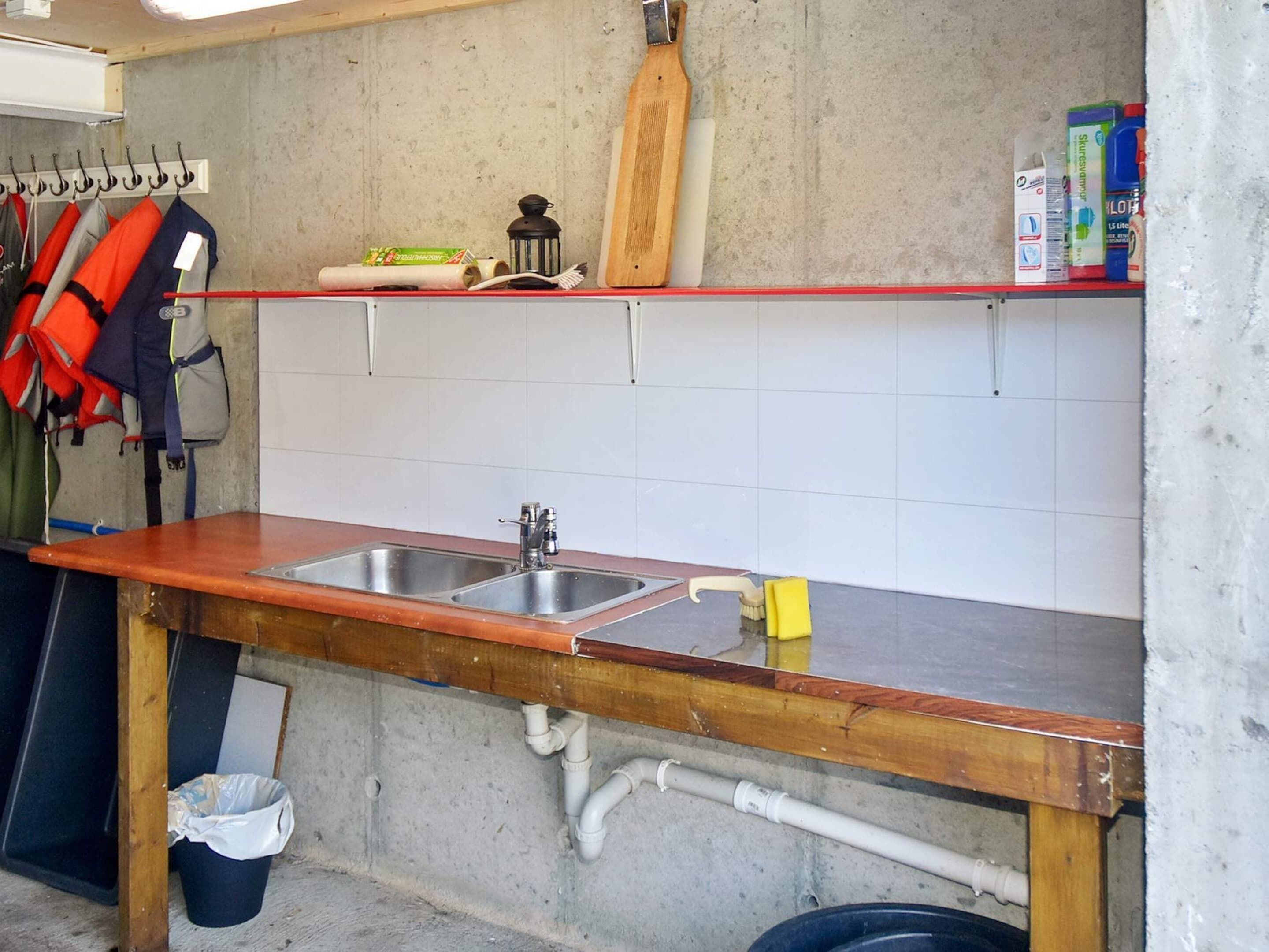 A kitchen with two sinks, a wooden countertop, a hanging rack with life jackets, and a shelf with cleaning supplies.