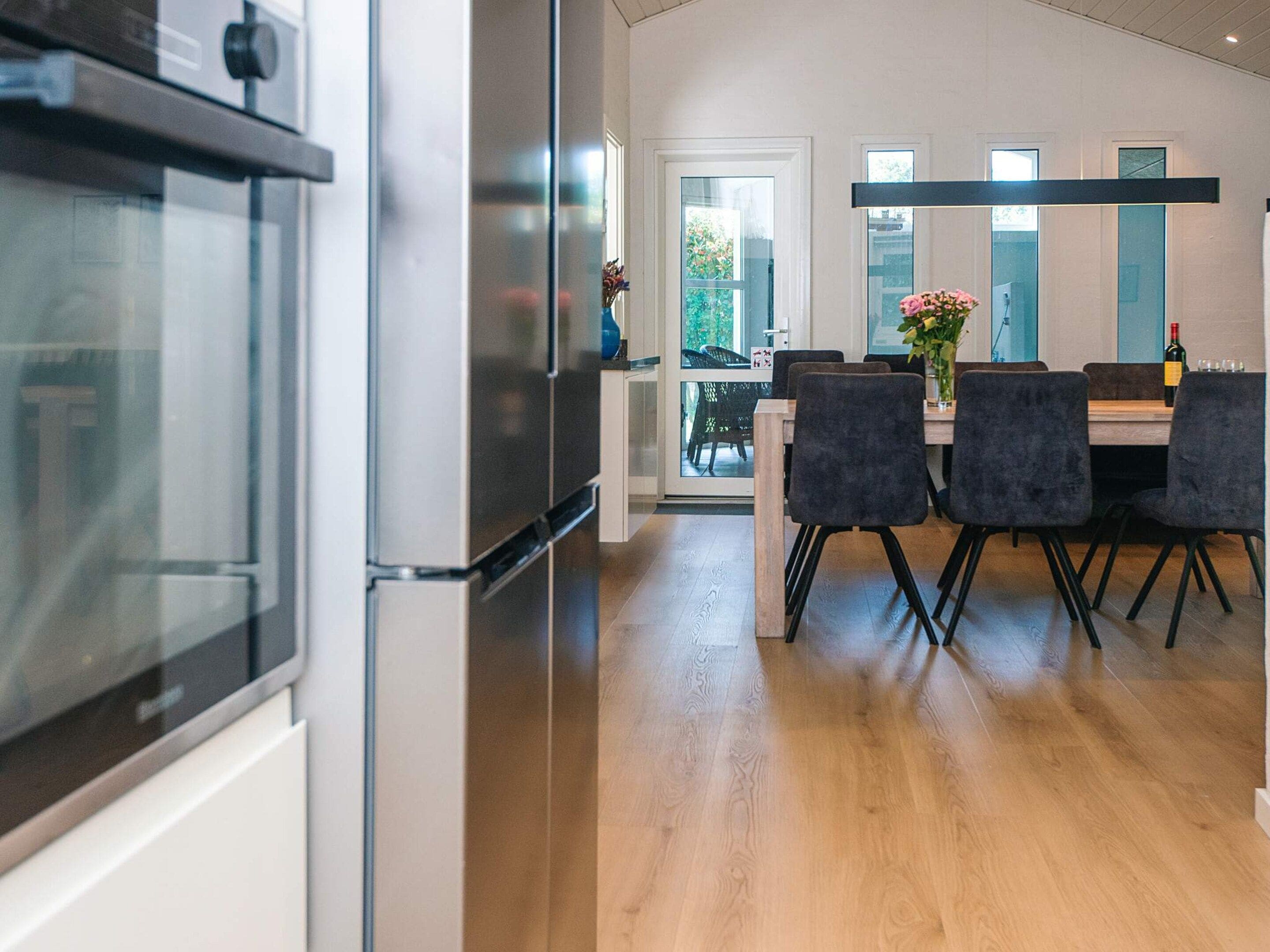 A modern kitchen with a stainless steel refrigerator, a dining area with a table and chairs, and a view of the outdoors through a glass door.