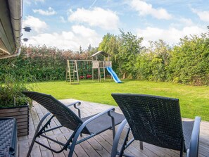 A backyard with a wooden play structure, including a slide, and two wicker chairs on a wooden deck. - 5 Star Holiday Home in Juelsminde (Horsens)