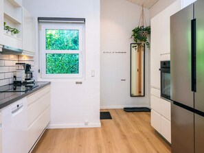 A modern kitchen with white cabinetry, a built-in oven, a stainless steel refrigerator, a coffee maker, and a window with a view of greenery. - 5 Star Holiday Home in Juelsminde (Horsens)