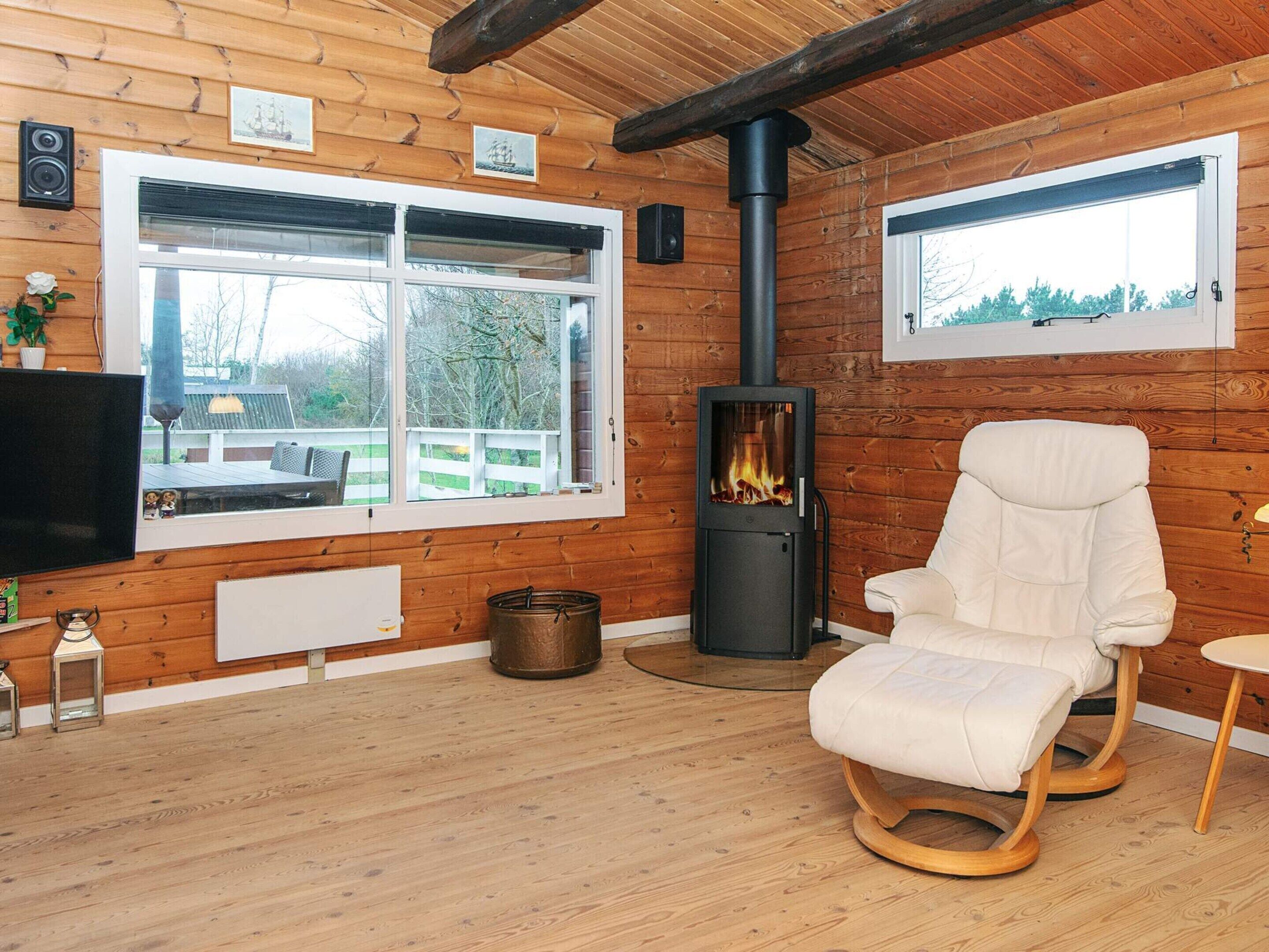 A cozy cabin interior with a large window, a wood-burning stove, a comfortable chair, and a television.