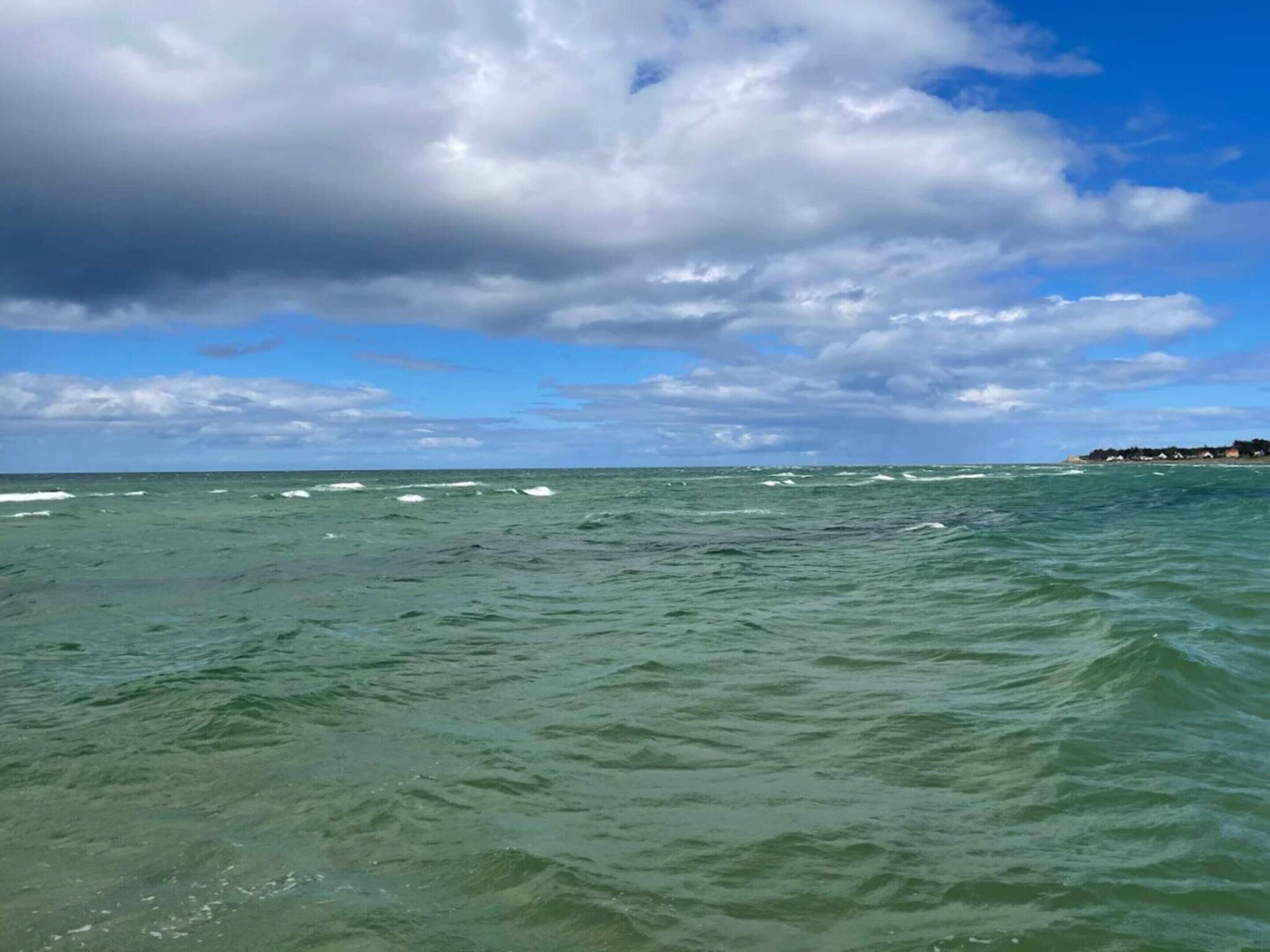 Blick auf die Küste mit Wellen, einem teilweise bewölkten Himmel und einer fernen Uferlinie mit Gebäuden.