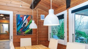 Een houten interieur met een eettafel, witte stoelen, een groot abstract schilderij, hanglampen en een raam met uitzicht op groen.