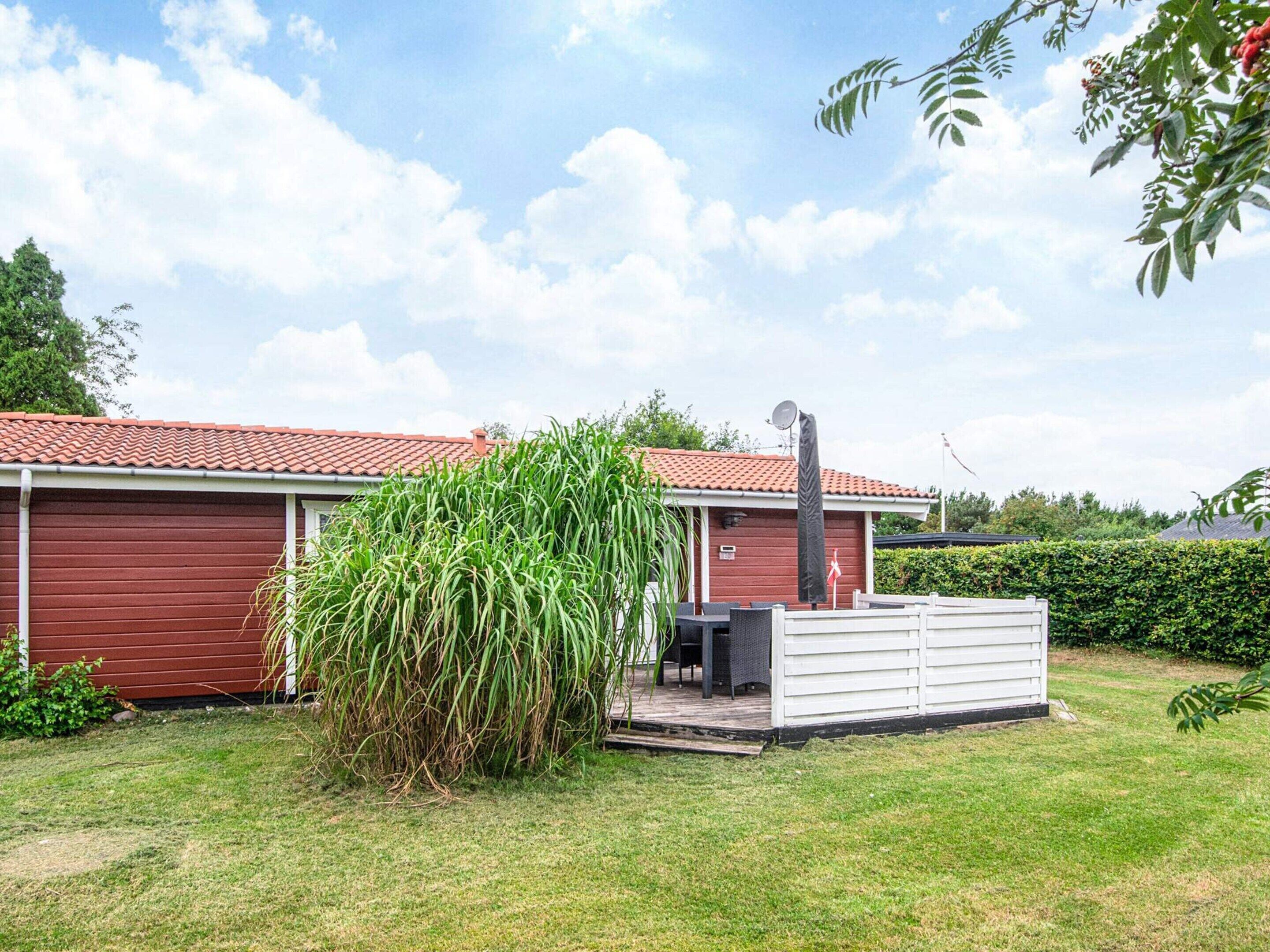 A red building with a tiled roof, a covered outdoor area with a grill, and a large bush in the foreground.