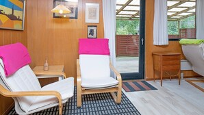 A room with wooden paneling, a bed with a pink blanket, a chair with a white cushion, a small table with a bottle, and a view through a glass door to an outdoor area. - 6 Person Holiday Home in Vaeggerlose-by Traum (Vaeggerlose)