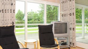 Une pièce avec deux fauteuils noirs rembourrés, une petite table ronde et un téléviseur posé sur un meuble. De grandes fenêtres offrent une vue sur la verdure environnante.