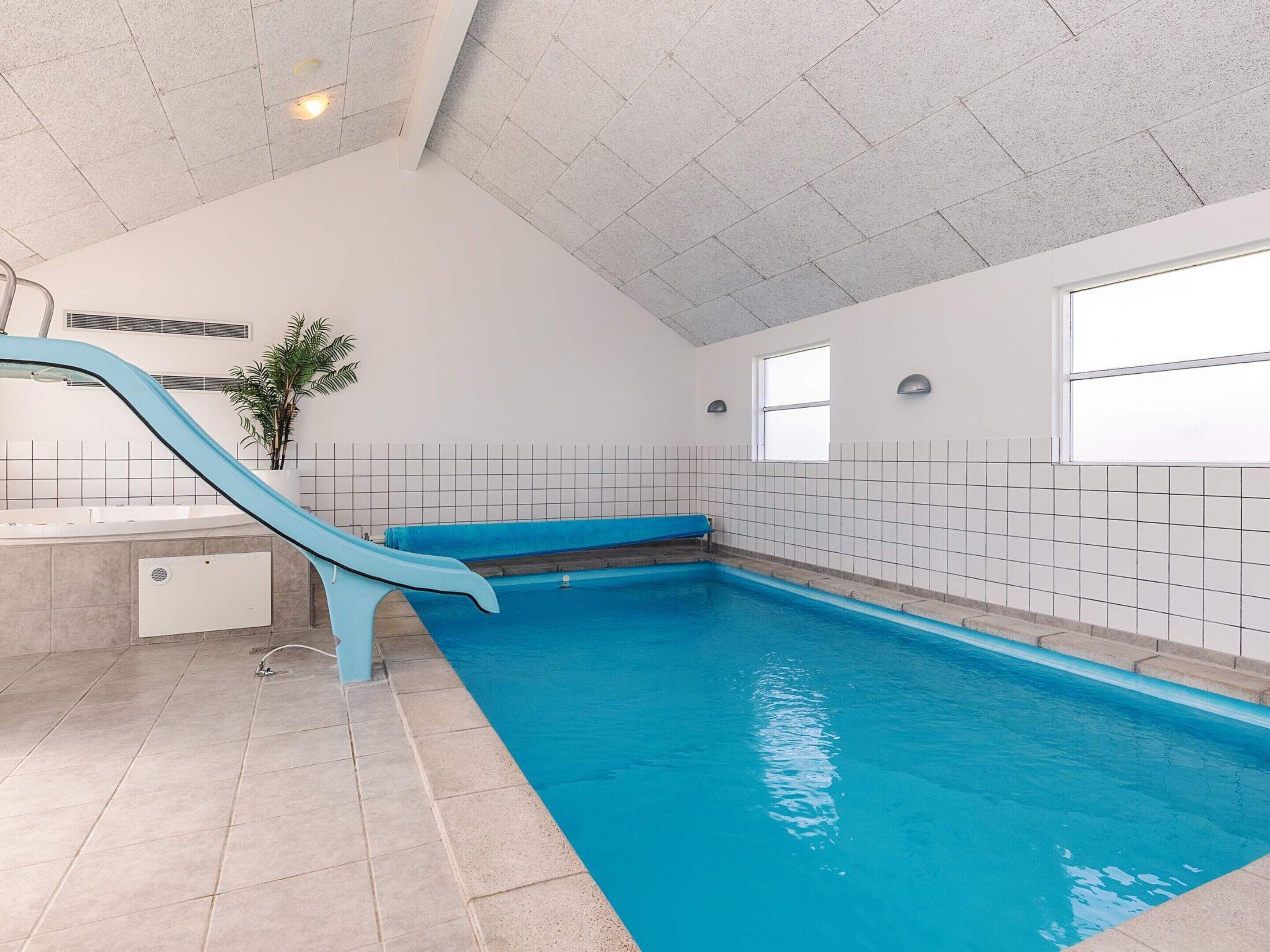 En indendørs swimmingpool med blå rutsjebane, flisebeklædte vægge og et loft med et gittermønster.