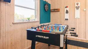 Une pièce aux murs en bois, avec une table de baby-foot, un jeu de fléchettes et une fenêtre ornée de plantes.