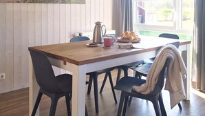 A dining area with a wooden table, black chairs, a coffee pot, a plate of fruit, and a window with curtains. - Holiday Home in Otterndorf (Otterndorf)