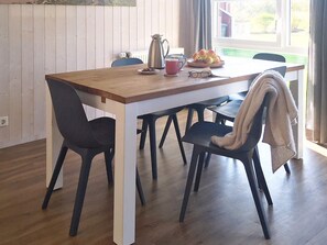 A dining area with a wooden table, black chairs, a coffee pot, a plate of fruit, and a window with curtains. - Holiday Home in Otterndorf (Otterndorf)