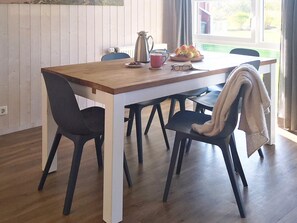 Un coin repas avec une table en bois, des chaises noires, une cafetière, une assiette de fruits et une fenêtre avec des rideaux.