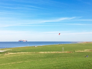 A grassy field with a body of water, a ship, and a kite in the sky. - Holiday Home in Otterndorf (Otterndorf)