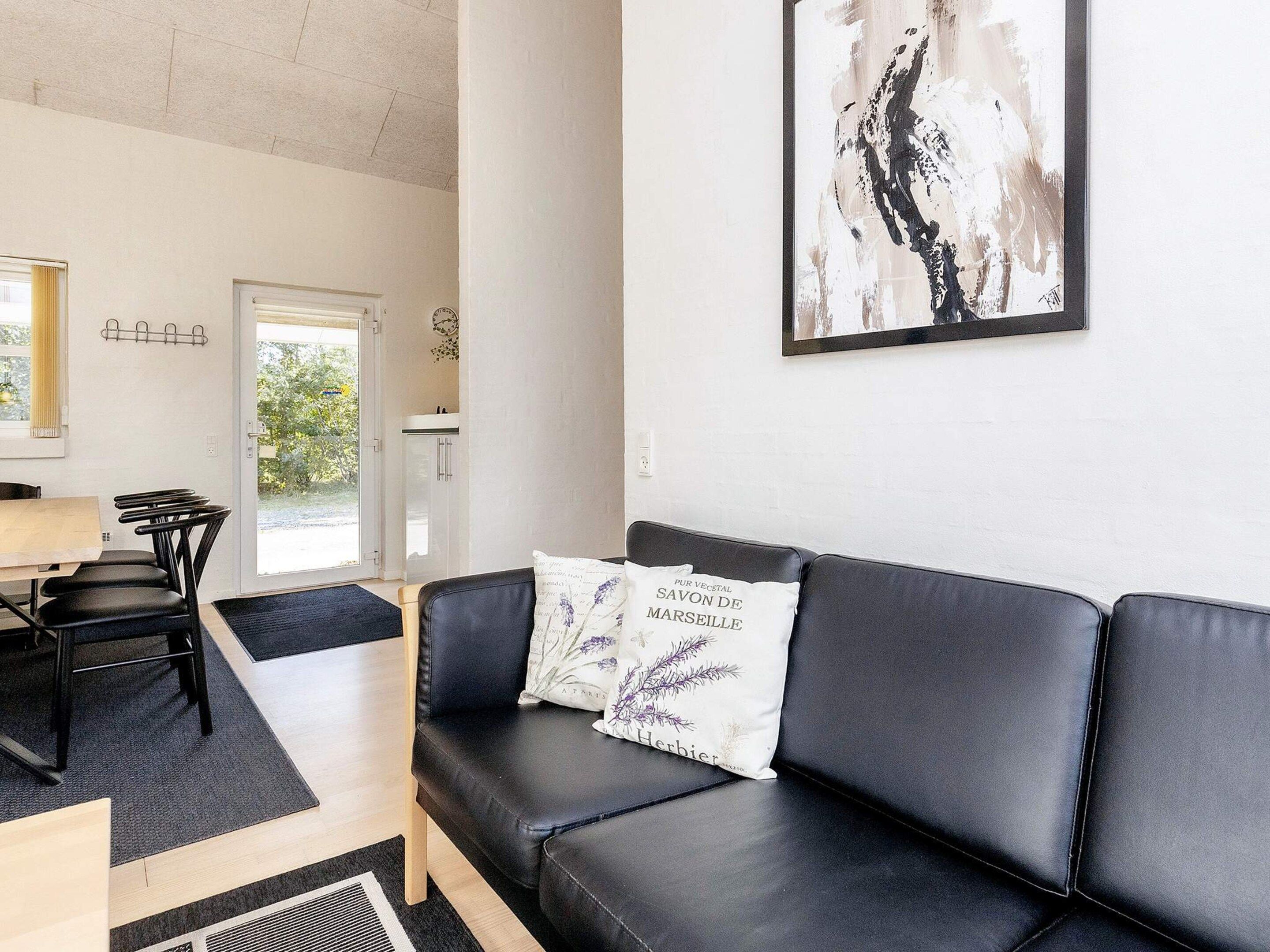 A modern living room with a black leather sofa, a dining table with chairs, and a kitchen area visible through an open door.