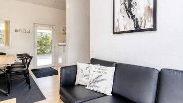 A modern living room with a black leather sofa, a dining table with chairs, and a kitchen area visible through an open door.