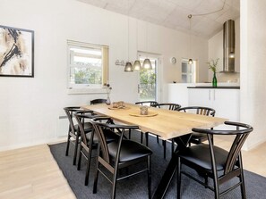 A dining area with a wooden table and black chairs, a window with curtains, and a kitchen in the background.