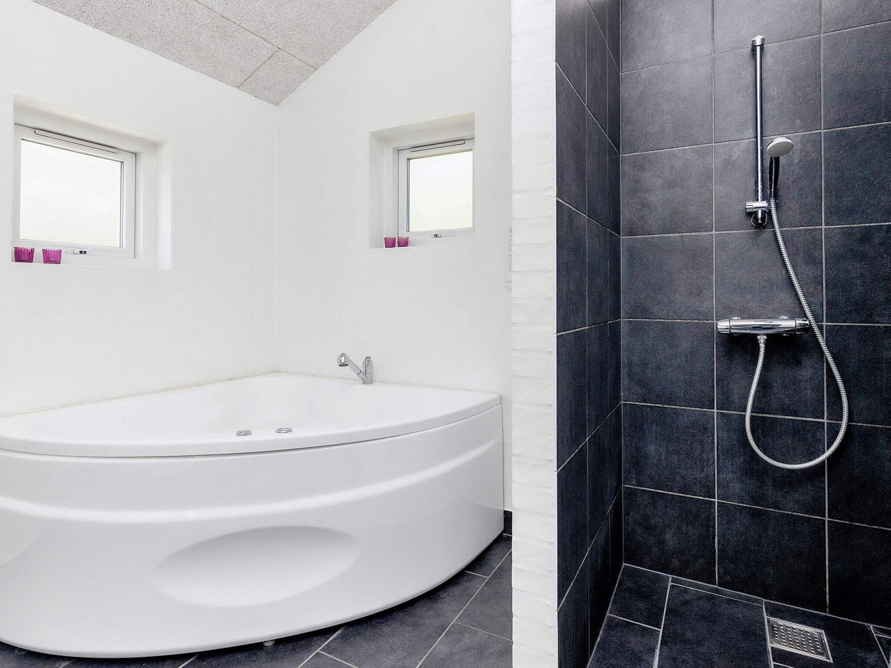 A modern bathroom with a freestanding bathtub, a shower with a handheld showerhead, and two windows with pink curtains.