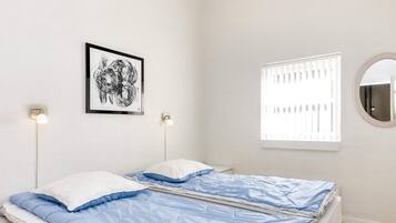 A bed with a blue and white duvet, two bedside lamps, a framed picture on the wall, a window with blinds, and a round mirror.