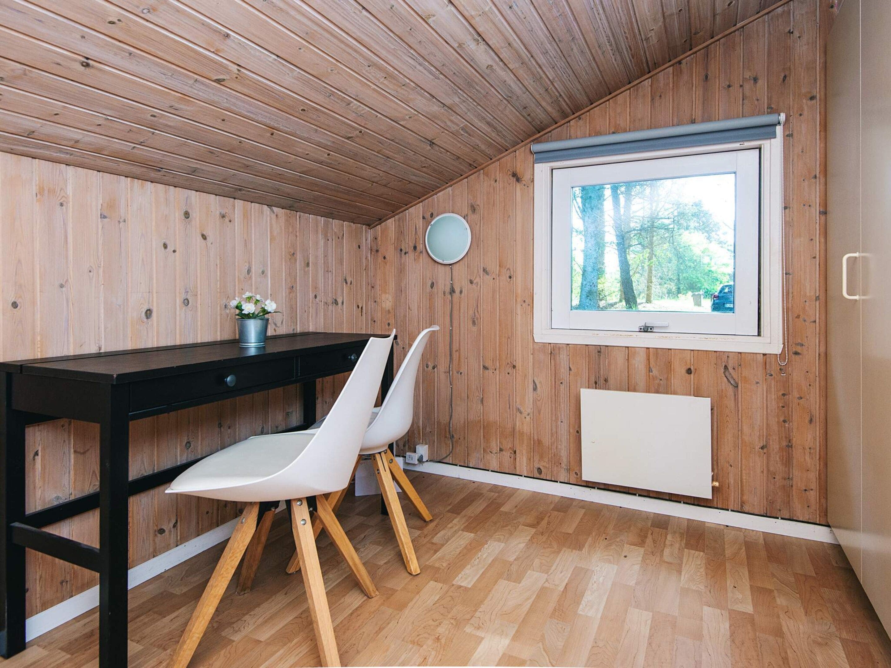 Ein kleiner Raum mit Holzvertäfelung und Holzboden, ein Fenster mit Blick auf Bäume, ein schwarzer Schreibtisch mit Stuhl und eine Heizung.