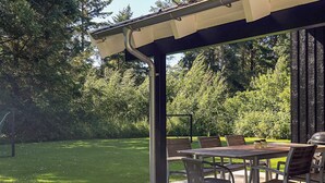 A covered outdoor patio with a table and chairs, surrounded by a grassy lawn and trees. - 4 Star Holiday Home in Bindslev (Bindslev)