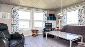 A cozy living room with a leather sofa, a black leather chair, a small wooden table, and a white table. There are two windows with curtains, a lamp, and a picture on the wall. - 6 Person Holiday Home in Ringkobing (Ringkobing)