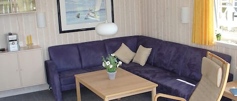 Una habitación con un sofá morado, una mesa de madera con un jarrón de flores, una silla, un cuadro en la pared y una ventana con cortinas amarillas.